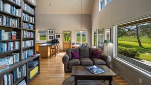 A spacious living room with a large bookshelf, a cozy sofa, and a dining area with a view of a green landscape. - Crow Cabin- Cozy Cabin with amazing views on 20 Acres in the Paradise Valley! (Livingston)