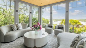 A modern living room with a curved gray sofa, two armchairs, and a round coffee table with a vase of flowers. Large windows offer a view of greenery and a body of water. - Ocean, golf and lagoon views, complete renovation luxury home (Kiawah Island)