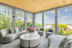 A modern living room with a curved gray sofa, two armchairs, and a round coffee table with a vase of flowers. Large windows offer a view of greenery and a body of water.