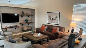 Een moderne woonkamer met een bruine leren bank, grijze fauteuils, een salontafel en een flatscreen televisie aan de muur.