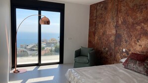 Une chambre à coucher avec un lit, un fauteuil vert, une lampe sur pied et une vue sur la mer par une grande fenêtre.