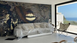Un salon moderne avec une grande fresque murale abstraite, un canapé recouvert d’un tissu, une chaise pliante et une vue sur l’océan.