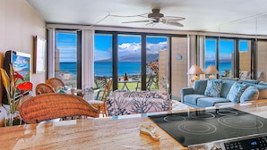 A spacious living room with a large window offering a view of the ocean, a ceiling fan, a dining area with a table and chairs, and a sofa with decorative pillows. - Kaleialoha 106 Ocean Front 1bd/1ba 1 Bedroom Condo by RedAwning (Lahaina)