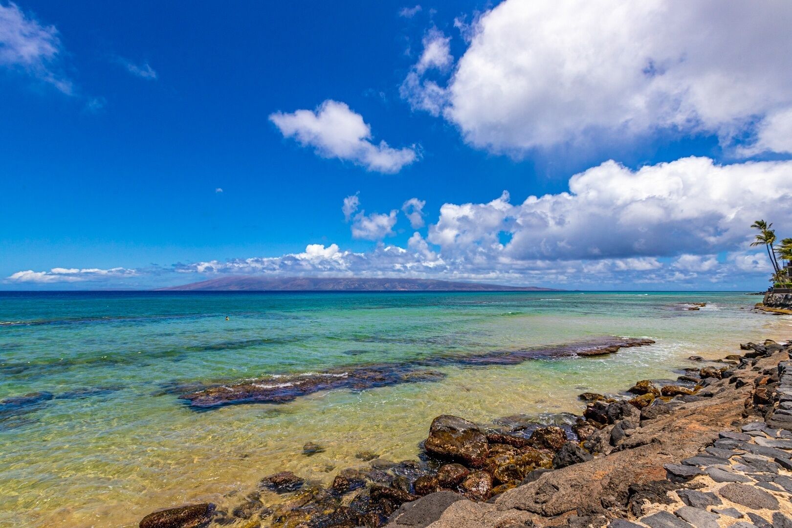 A clear view of a rocky shoreline with calm turquoise waters, a distant mountain range, and a partly cloudy sky.