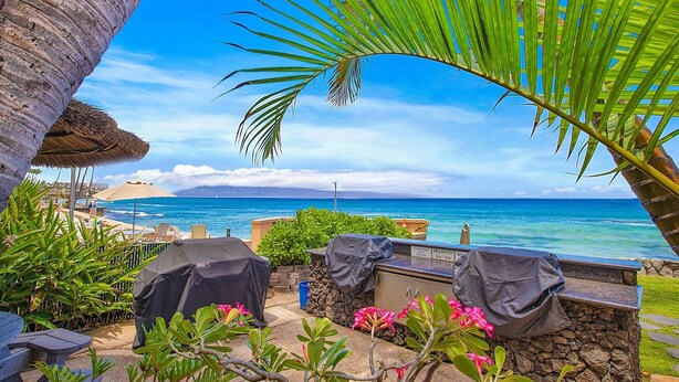 A patio area with a view of the ocean, covered barbecue grills, and a thatched umbrella. - Kaleialoha 106 Ocean Front 1bd/1ba 1 Bedroom Condo by RedAwning (Lahaina)