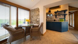 A kitchen with a stone wall, blue cabinets, and a dining area with wicker chairs and a glass table. - Charming countryside cottage for couples with private hot tub and communal games room. (Barnstaple)