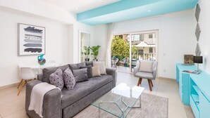Ein modernes Wohnzimmer mit grauem Sofa, einem Glastisch und einem Deckenventilator. Durch das Fenster ist ein Balkon mit Gartenmöbeln zu sehen.