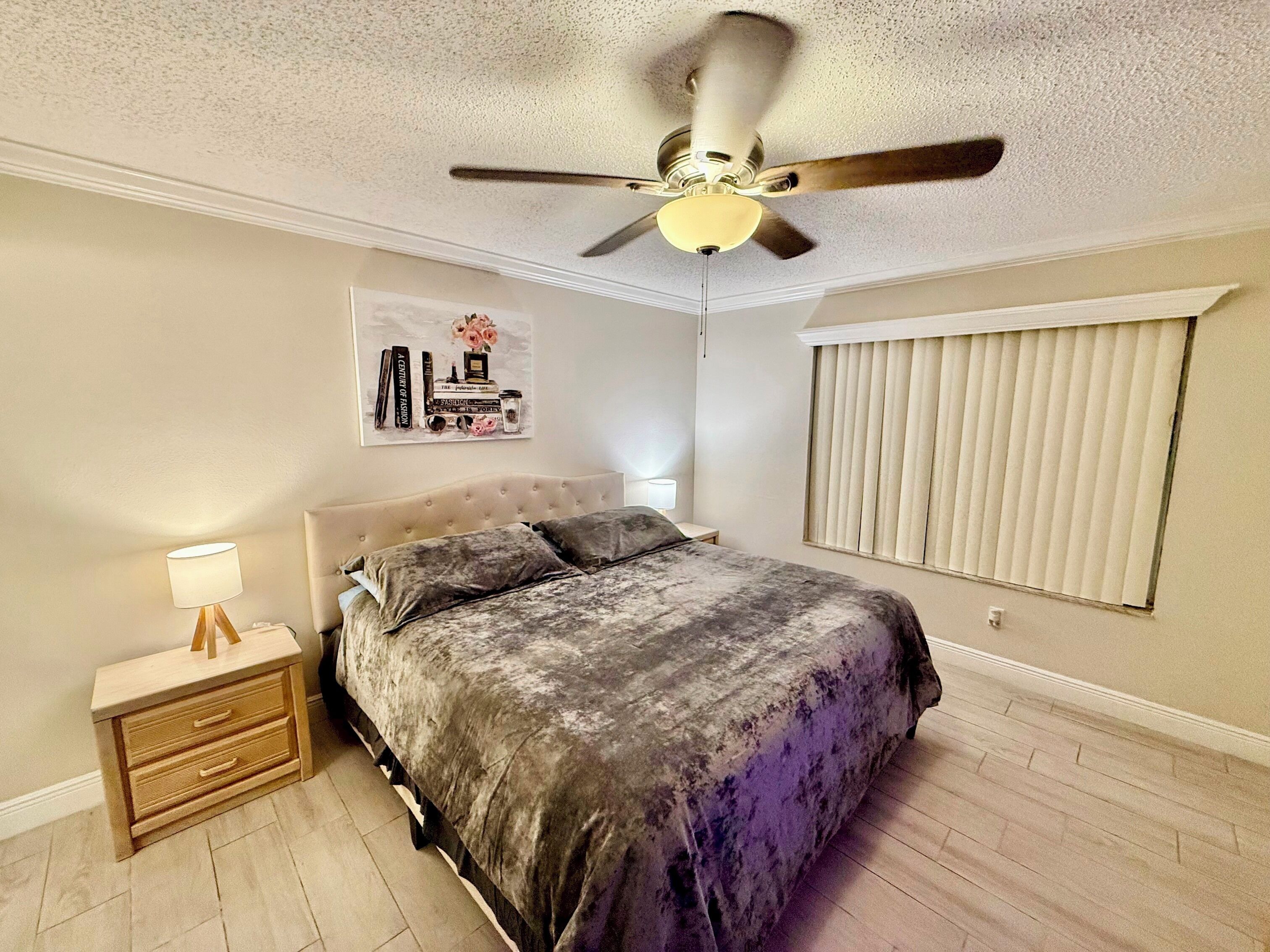 Un dormitorio con cama, mesita de noche, lámpara, ventilador de techo y una ventana con persianas.