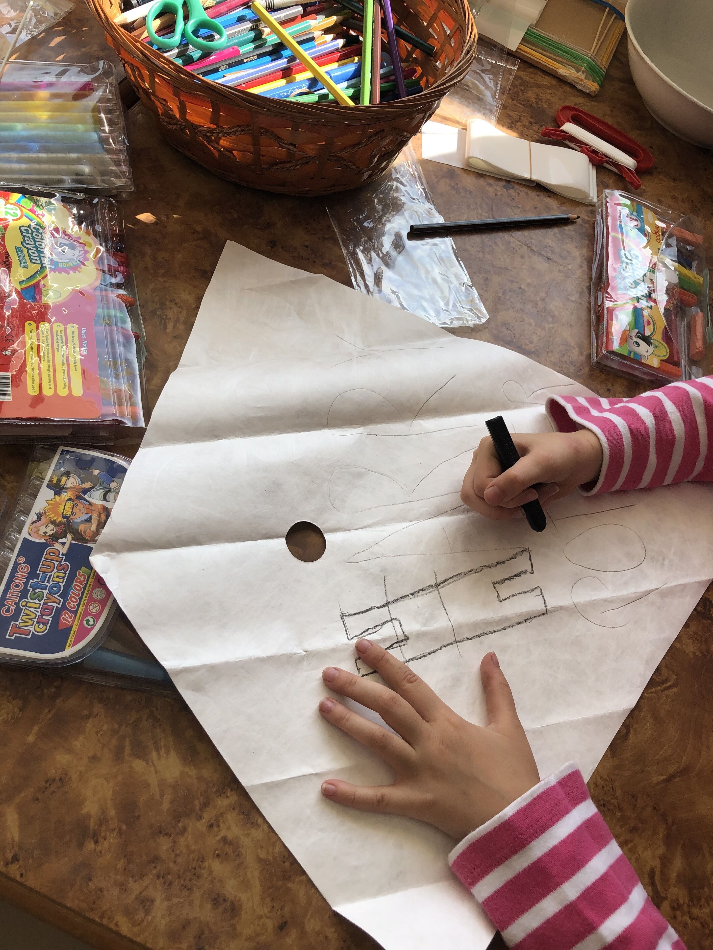 A person drawing on a large sheet of paper with a black marker, surrounded by various art supplies including crayons, pencils, and scissors on a wooden table.
