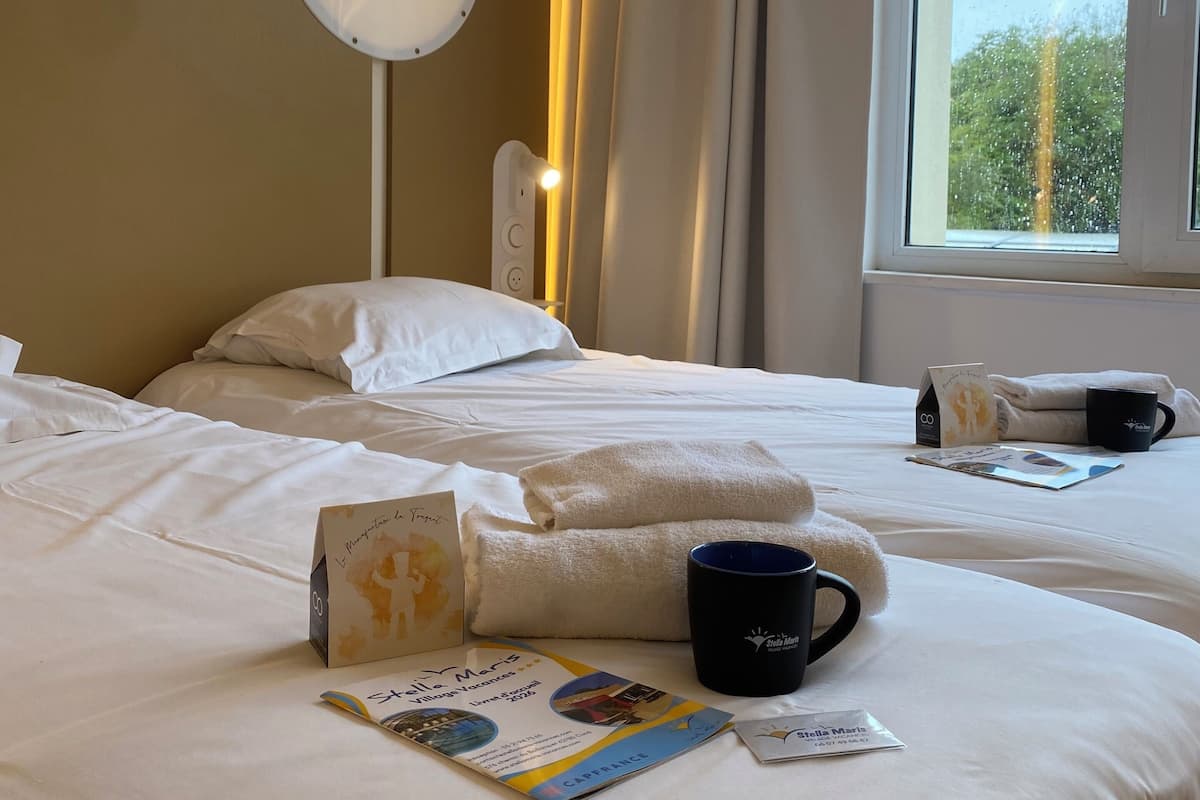A hotel room with two beds, a bedside table with a book and a mug, a mirror, and a window with curtains.