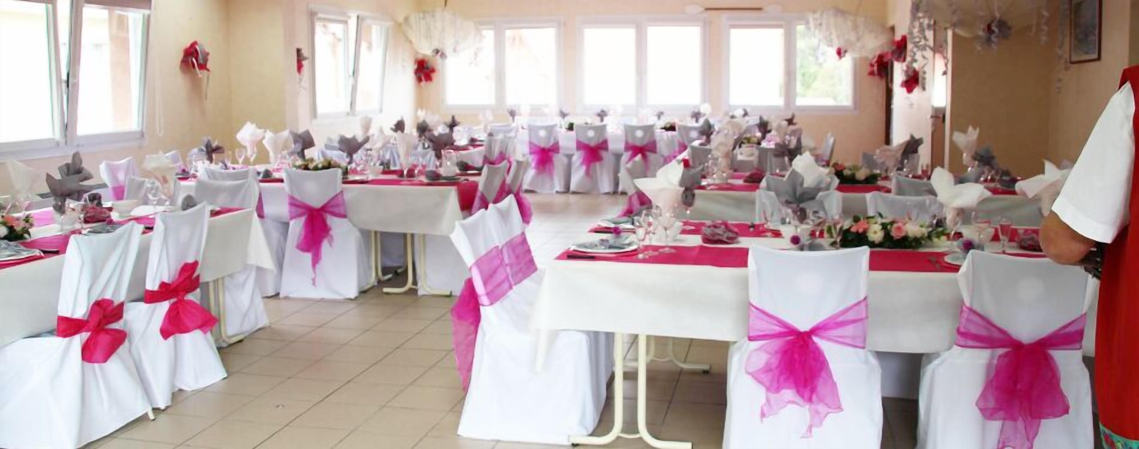 Une salle de banquet où les tables sont dressées pour un événement formel, avec des nappes blanches et des chaises ornées de rubans roses.