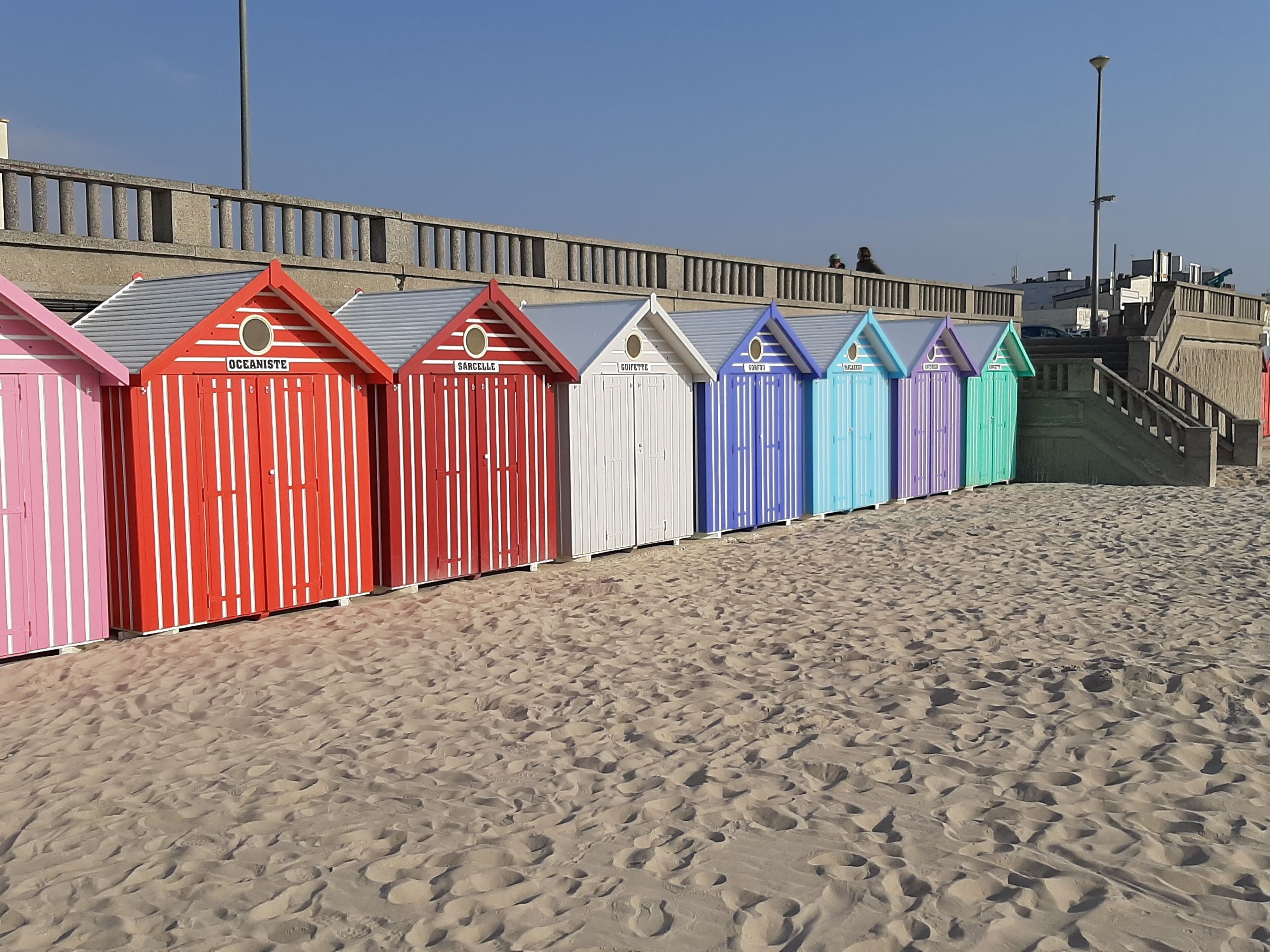Eine Reihe von Strandhütten mit Namen wie 'OCEANISTE' und 'SARCELLE' an einem sandigen Strand.