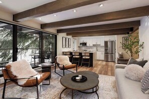 A modern kitchen with stainless steel appliances, a dining area with leather chairs, and a large window offering a view of trees.