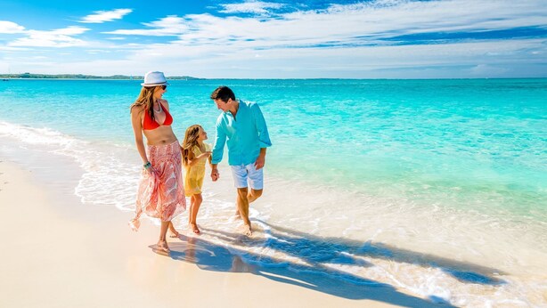 Une famille se promène sur une plage où l’eau est d’un bleu turquoise limpide et le ciel est dégagé.