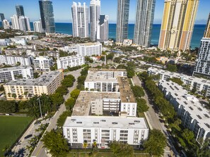 A high-rise building with a flat roof and multiple floors, surrounded by other buildings and a body of water in the background. - Walk to the Beach at Sunny Isles (Sunny Isles Beach)