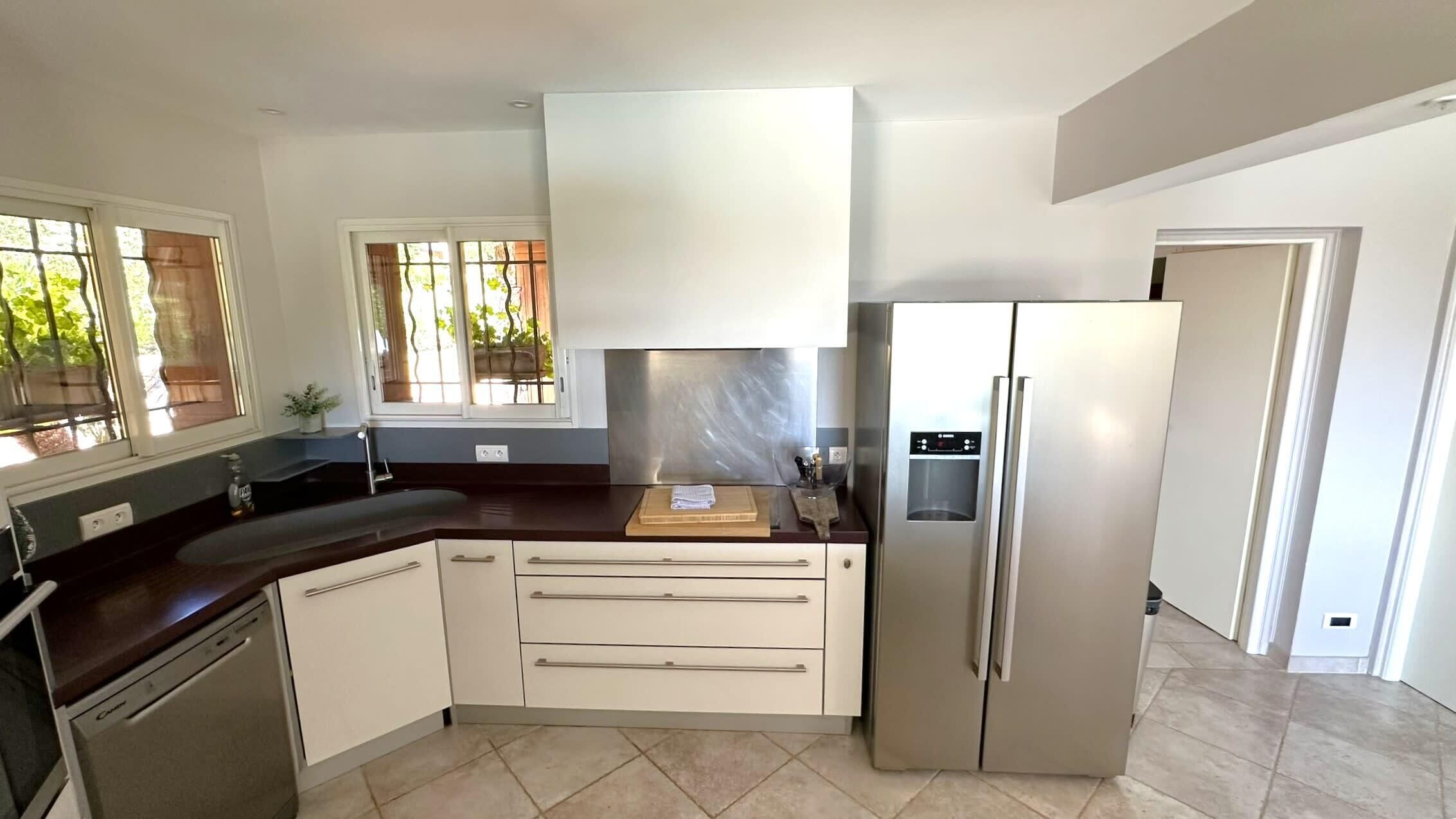 Une cuisine moderne avec des meubles blancs, un réfrigérateur en acier inoxydable, un four encastré et une fenêtre donnant sur un paysage verdoyant.