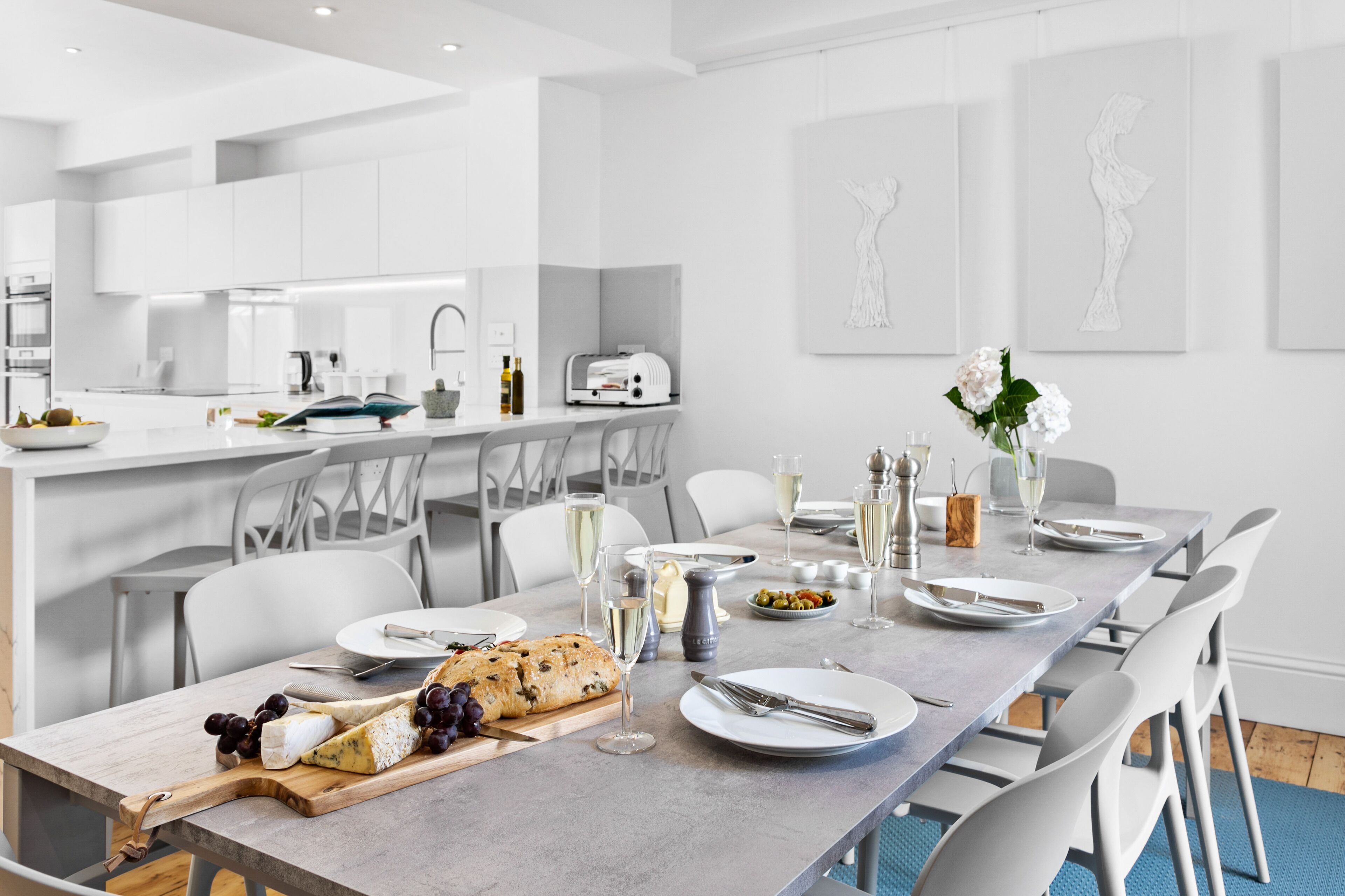 A modern dining area with a long table set for a meal, white chairs, and a kitchen in the background.