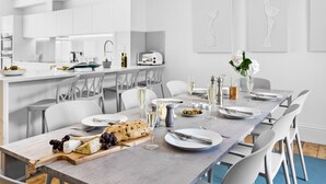 Ein moderner Essbereich mit einem langen Tisch, der für eine Mahlzeit gedeckt ist, weißen Stühlen und einer Küche im Hintergrund.