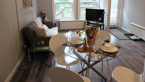 A dining area with a glass table set for four, surrounded by white chairs. A piano is visible in the background, and a window with a view of greenery is present.