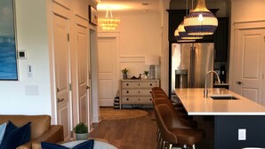 Una cuina moderna amb una illa central, tamborets, llums penjants i una zona de menjador amb una cadira rodona i coixins.