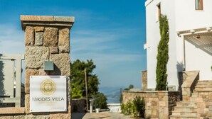A sign for TZIKIDES VILLA, a luxury villa for rent, with a stone wall and a view of a white building and greenery in the background. - Villa Tzikides Aegina (Aegina)