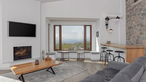 A modern living room with a fireplace, a large window with a view, a bar area, and a grey sofa. - Villa Tzikides Aegina (Aegina)