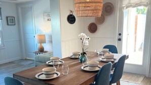 Un coin repas avec une table en bois dressée pour six personnes, des chaises bleues, un luminaire suspendu en rotin et une vue sur l’extérieur par une porte.