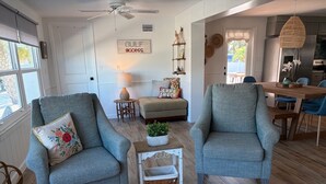 Un salon comprenant deux fauteuils, une table basse, un tapis, un coin repas avec une table et des chaises, et une cuisine visible à travers une porte ouverte.