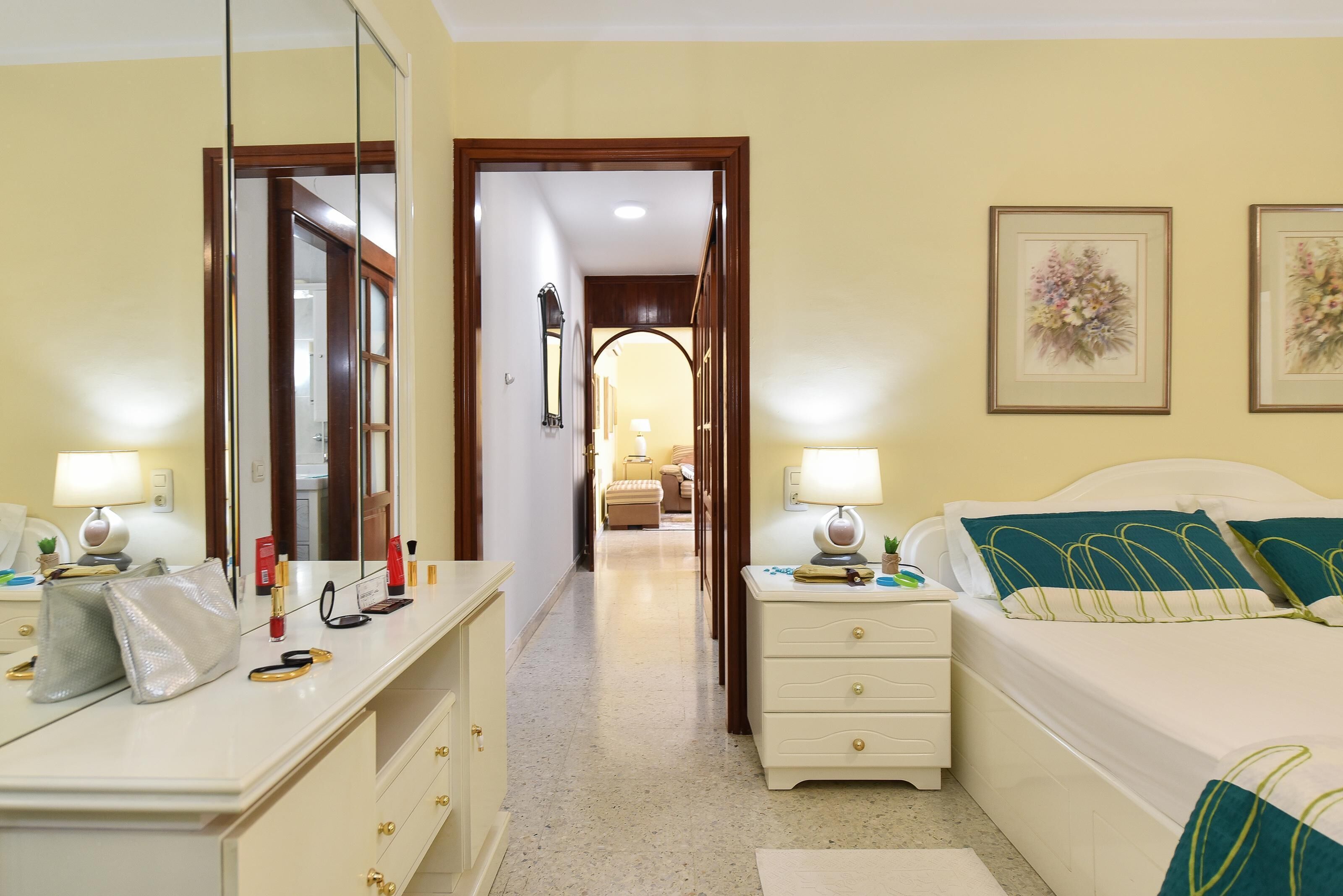 Ett hotellrum med en säng, sängbord, en byrå, en spegel och en hall som leder till ett annat rum.