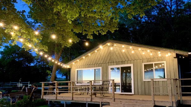A wooden cabin with a deck, string lights, and a large window.