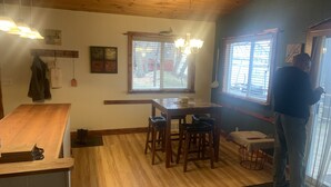 A wooden interior with a dining table, chairs, a bench, and a person standing by a window.