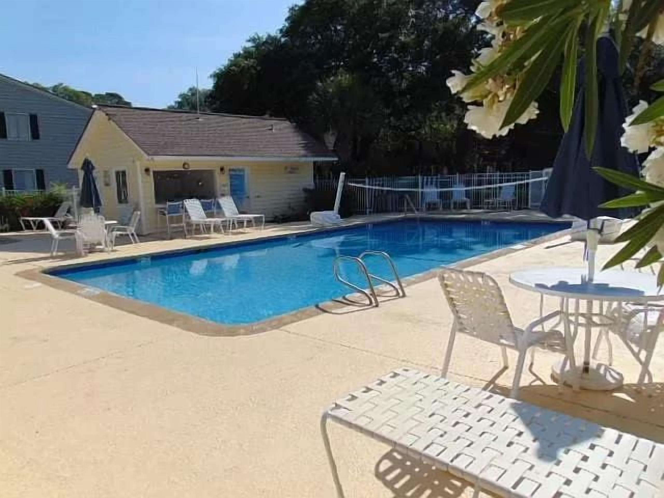Ein Pool mit Liegestühlen und ein kleines Gebäude, vermutlich ein Poolhaus, in einer sonnigen Umgebung im Freien.