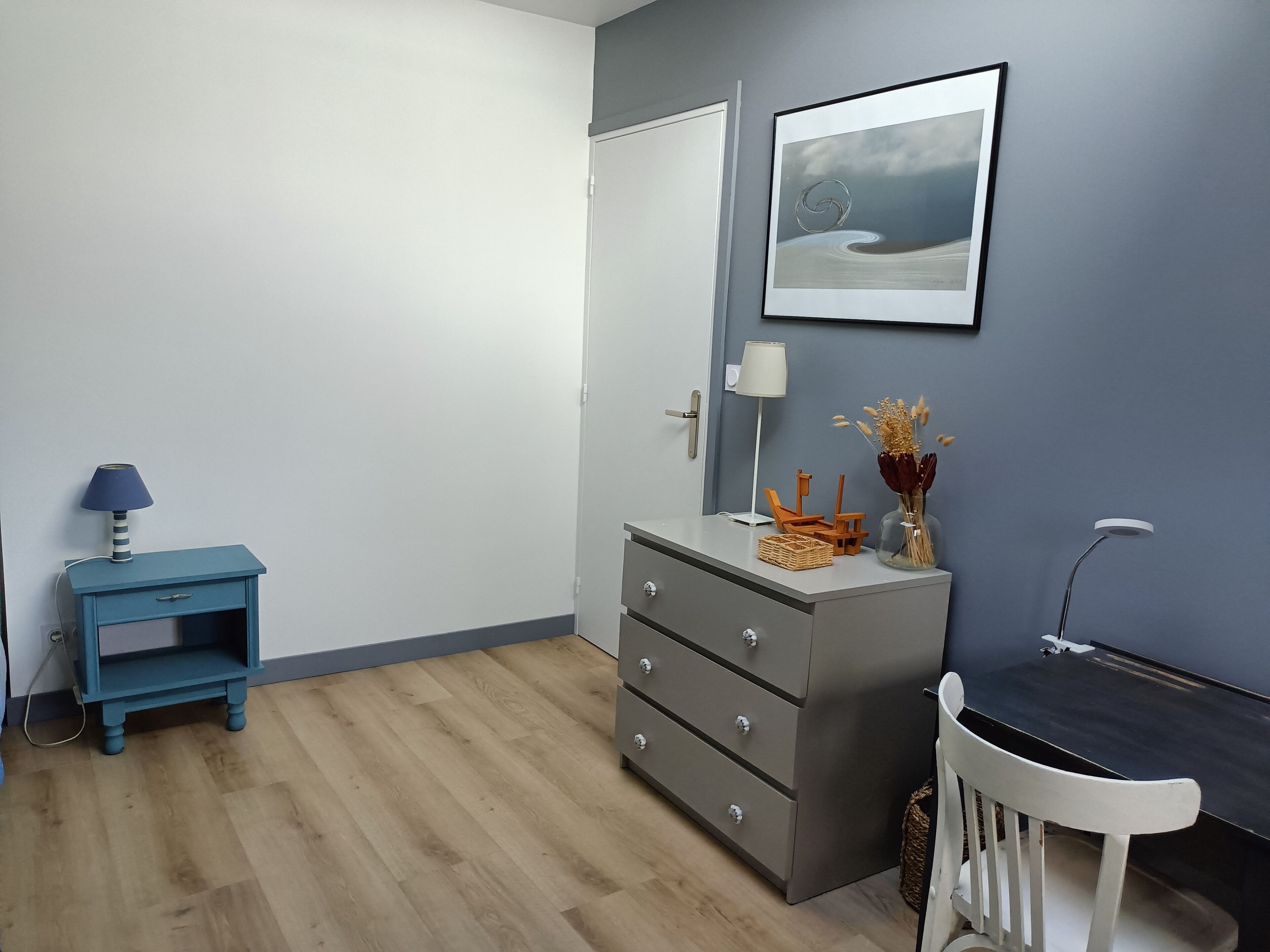 A room with a grey dresser, a small blue nightstand with a lamp, a white chair, and a framed picture on the wall.