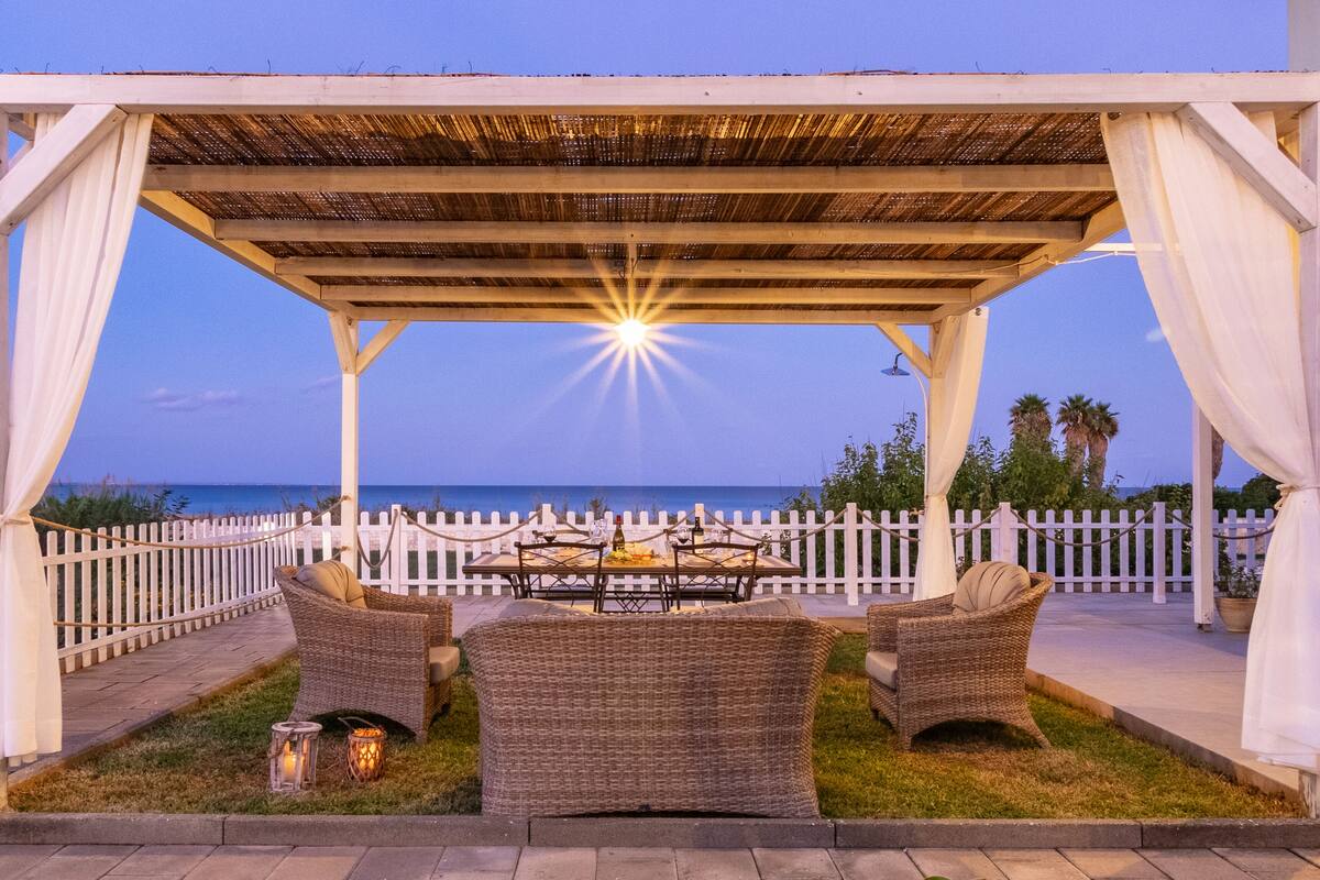 An outdoor seating area with wicker furniture and a fire pit, overlooking a white picket fence and the ocean.