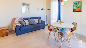 Un salon chaleureux avec un canapé bleu, une table de salle à manger ronde, des chaises colorées, un lampadaire et des œuvres d’art accrochées aux murs.