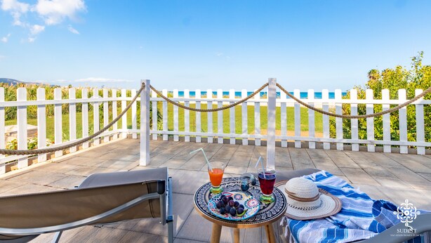 Une terrasse avec une table dressée pour deux, agrémentée de boissons et d’une assiette de fruits, donnant sur une clôture blanche et un ciel bleu dégagé.