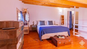 Une chambre avec un plafond en bois, un lit recouvert d’une couverture bleue, une commode en bois, une table de chevet et une valise posée sur le sol.