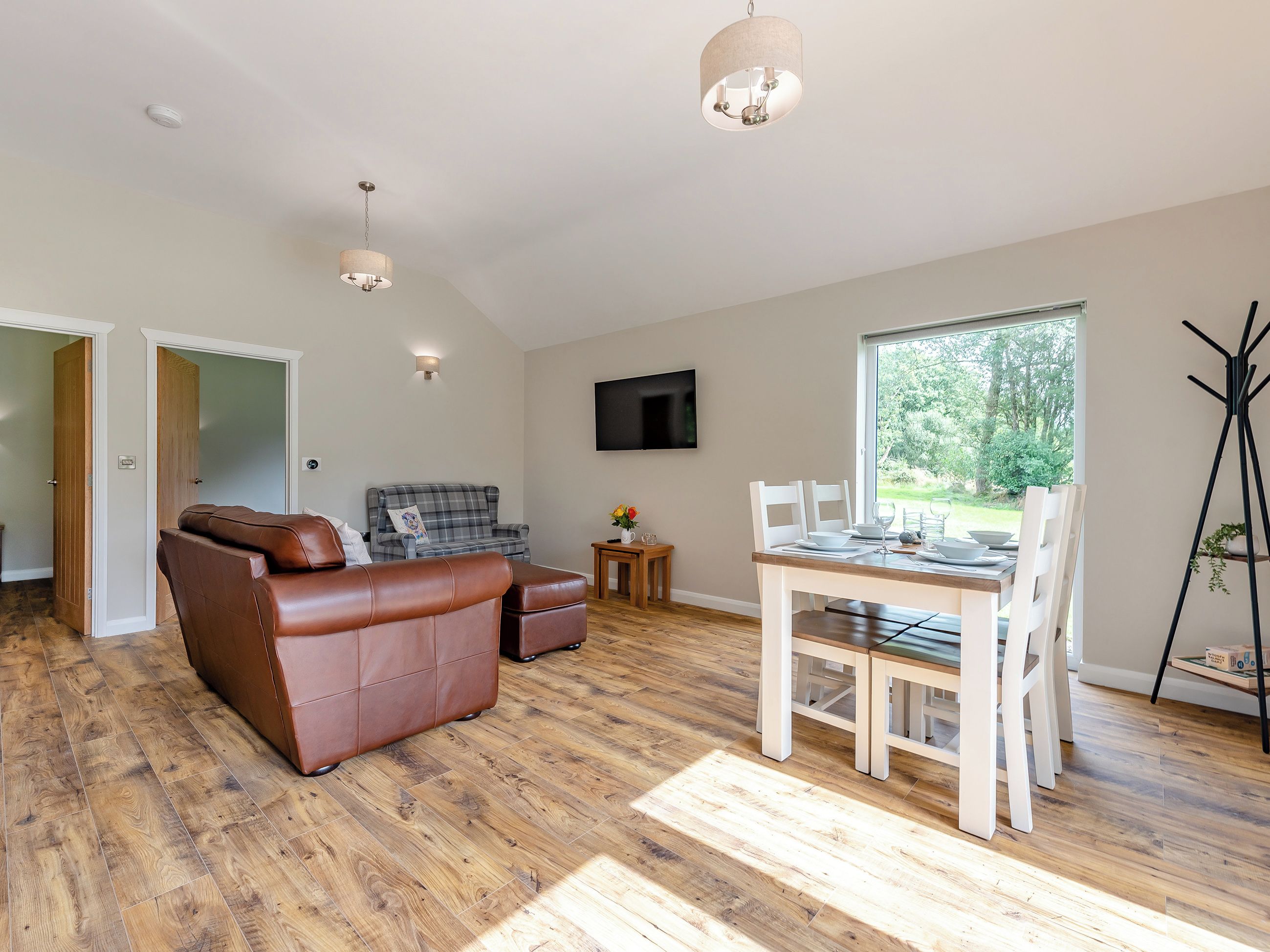 A spacious living room with a brown leather sofa, a plaid sofa, a dining table with white chairs, and a large window offering a view of greenery.
