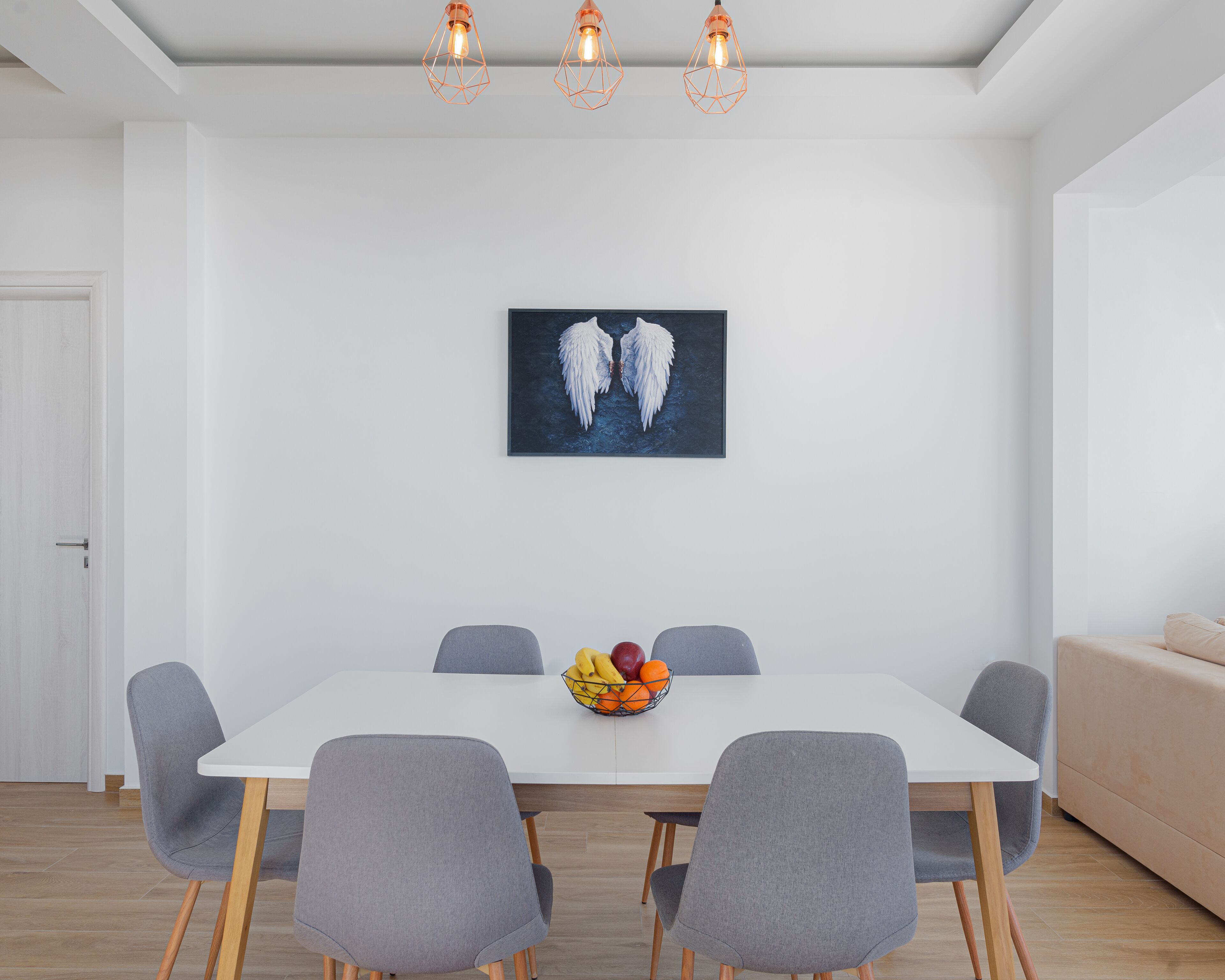 A dining area with a white table, four grey chairs, a fruit bowl, and a framed artwork of angel wings on the wall.