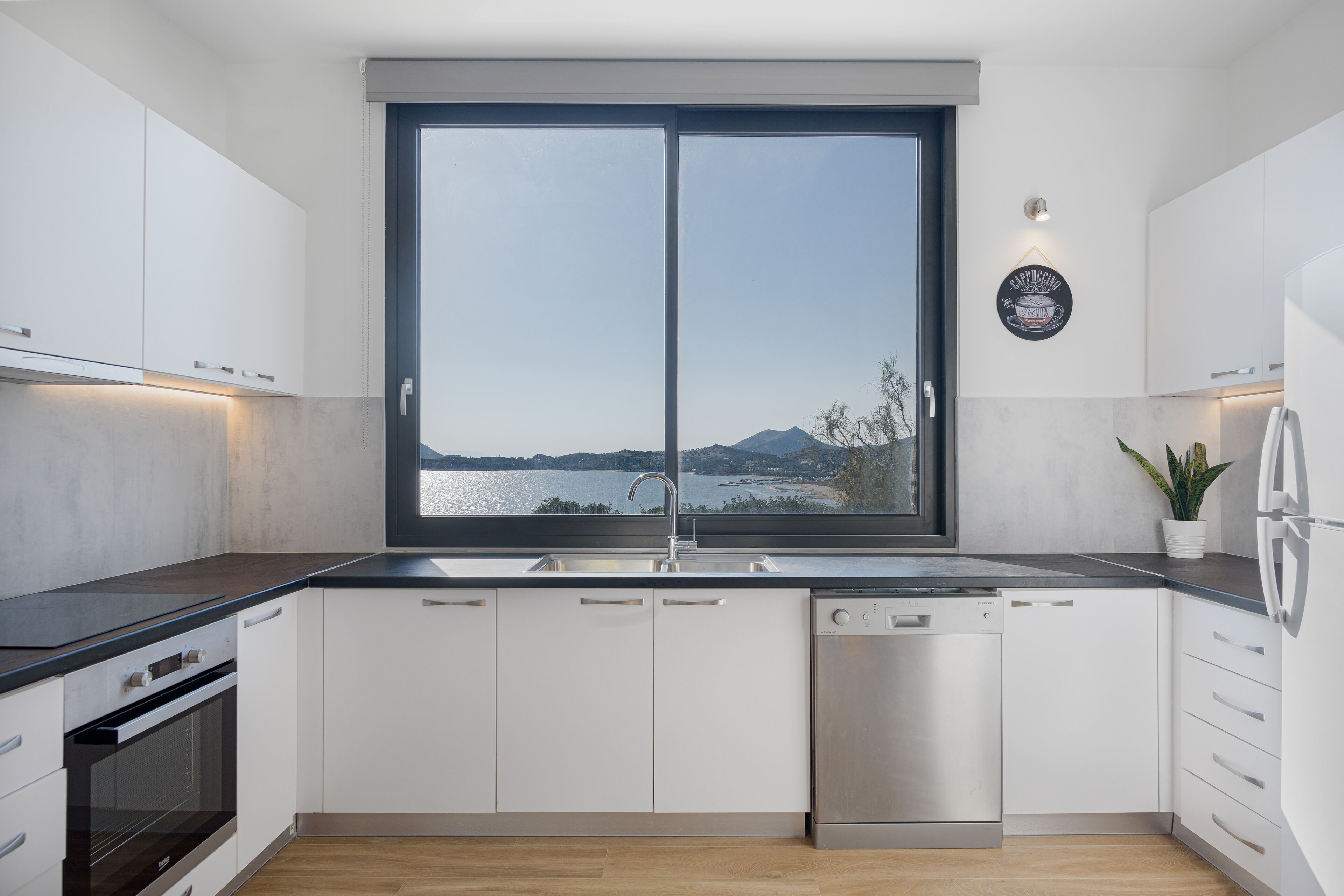 A modern kitchen with white cabinets, a black countertop, a dishwasher, and a large window offering a view of a lake and mountains.