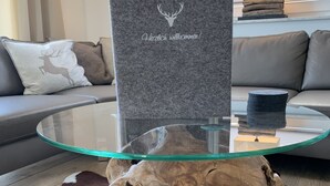 Une table basse avec un plateau en verre, ornée d’un logo représentant un cerf et d’une inscription, un canapé en cuir avec un coussin à motif de cerf, et un grand piédestal en bois brut.