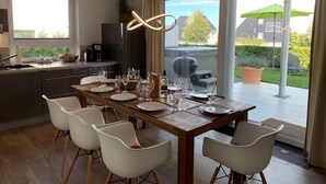 Un espace repas moderne avec une table en bois dressée pour six personnes, des chaises blanches et une vue sur le jardin à travers de grandes portes-fenêtres coulissantes.