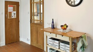 Un pasillo con una puerta de madera, un tablón de anuncios, un reloj de pared, una mesita con fruteros y cajas de almacenamiento.