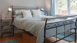 Una cama bien hecha con estructura metálica, una mesita de noche con lámpara y una ventana con cortinas.