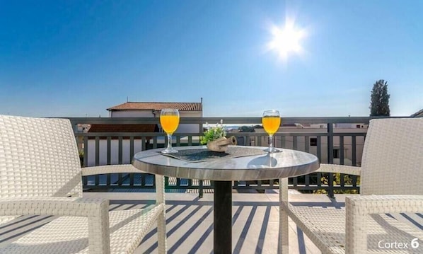 A round table with two glasses of orange juice, a small decorative item, and wicker chairs on an outdoor terrace with a clear sky and buildings in the background.