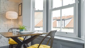 Un coin repas avec une table dressée pour deux personnes, une plante en pot et une vue sur le paysage urbain à travers de grandes fenêtres.