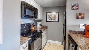A kitchen with white cabinets, a black stove, a microwave, and a refrigerator. There are two framed pictures on the wall. - Cozy Beach Condo with Private Beach, heated pool, High Speed WiFi, and More. (Orange Beach)
