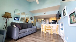 A living room with a grey sofa, wooden dining table, and chairs, a ceiling fan, and a kitchen area in the background. - Cozy Beach Condo with Private Beach, heated pool, High Speed WiFi, and More. (Orange Beach)