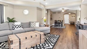 Ein modernes Wohnzimmer mit grauem Sofa, Couchtisch aus Holz und einem gemusterten Teppich. Es gibt einen Essbereich mit Tisch und Stühlen, sowie eine Küchenzeile mit Arbeitsfläche und Sitzgelegenheiten.