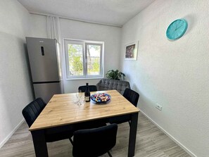 Cozinha compacta com mesa de madeira, cadeiras pretas, sofá cinza, geladeira, janela com vista e um relógio na parede.
